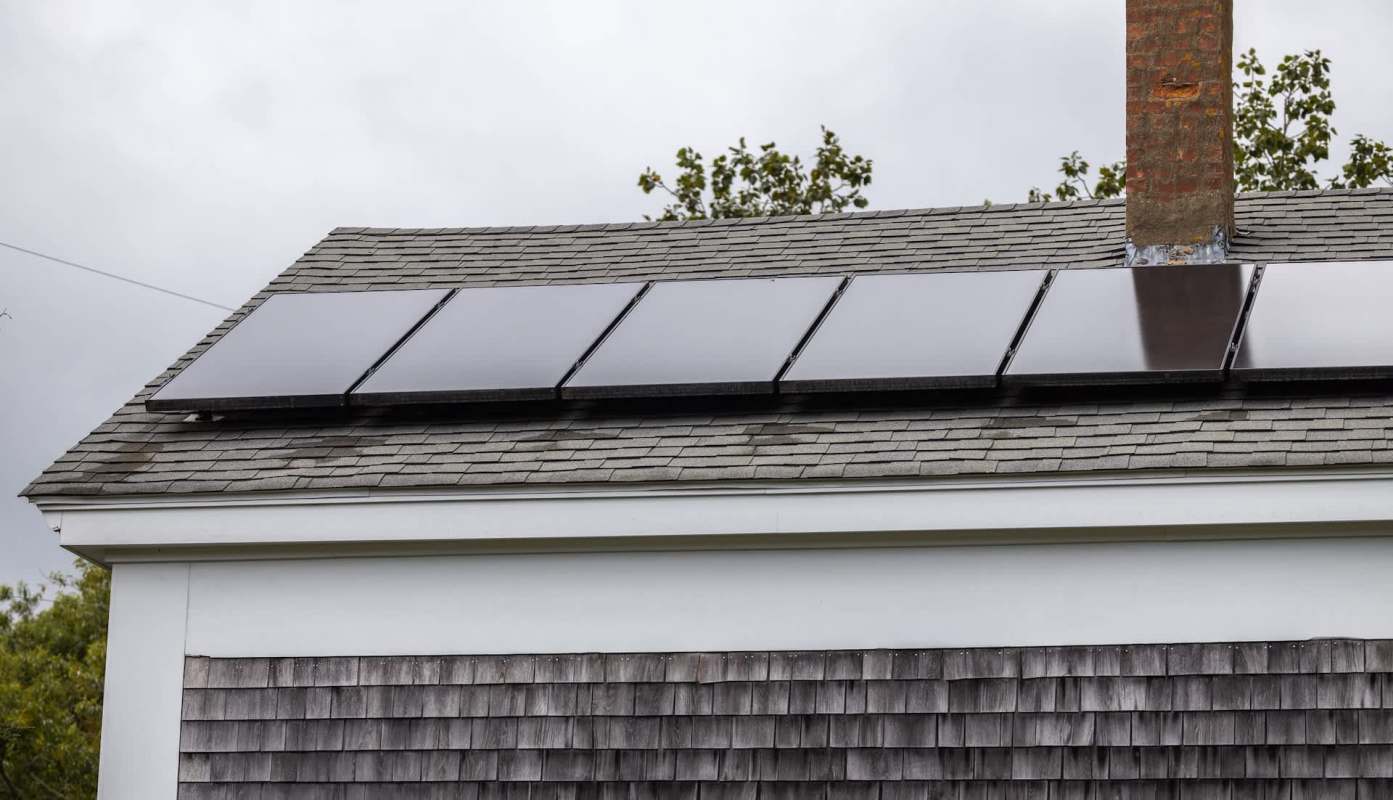 A roof with solar panels installed beside a brick chimney and wooden shingles.