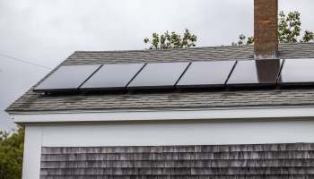 A roof with solar panels installed beside a brick chimney and wooden shingles.