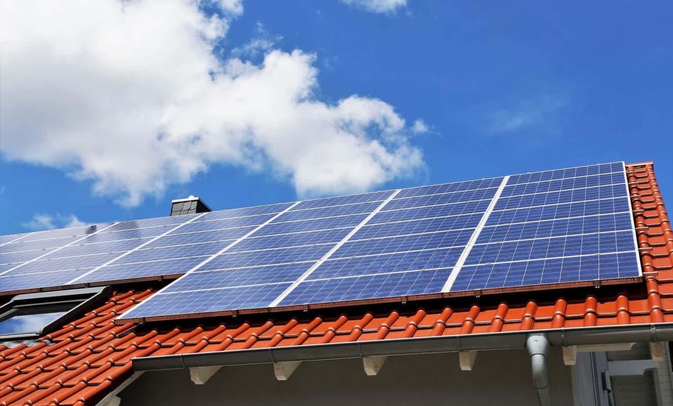 A rooftop with solar panels under a clear blue sky adorned with a few fluffy clouds.