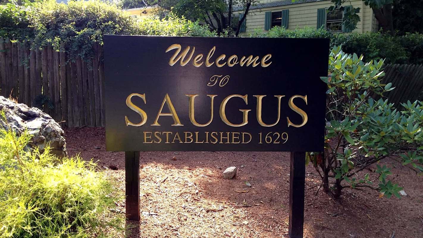 A welcome sign for Saugus, established in 1629, surrounded by greenery and wooden fencing.
