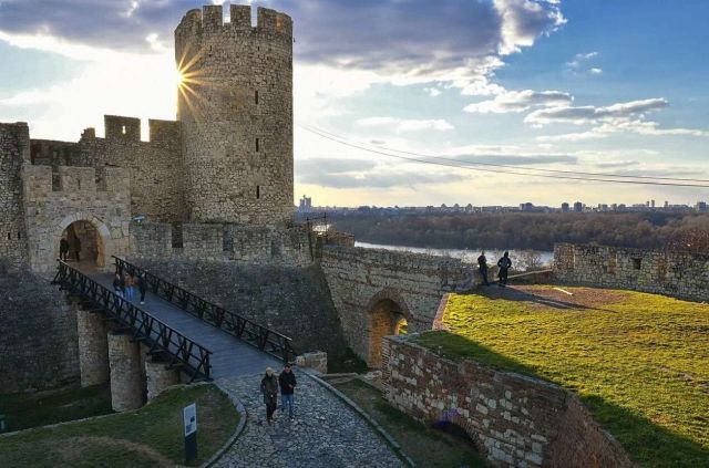 A historic stone castle with towers and a wooden bridge, overlooking a river and a cityscape in the background.