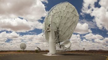 A large satellite dish stands on a desert landscape under a cloudy blue sky.