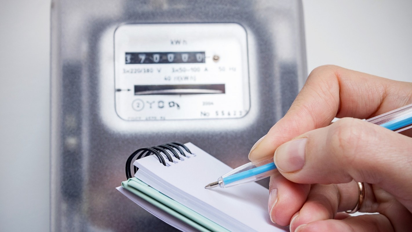 A hand holds a pen poised over a notebook, next to an electric meter displaying consumption readings.