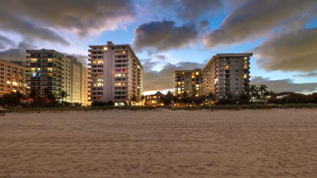 A beach view at dusk featuring tall buildings with illuminated windows and dramatic clouds in the sky.