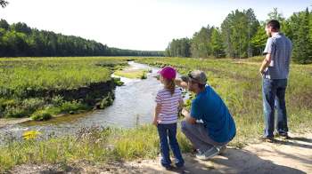 A child wearing a pink hat points at a river while an adult crouches beside her and another adult stands nearby.