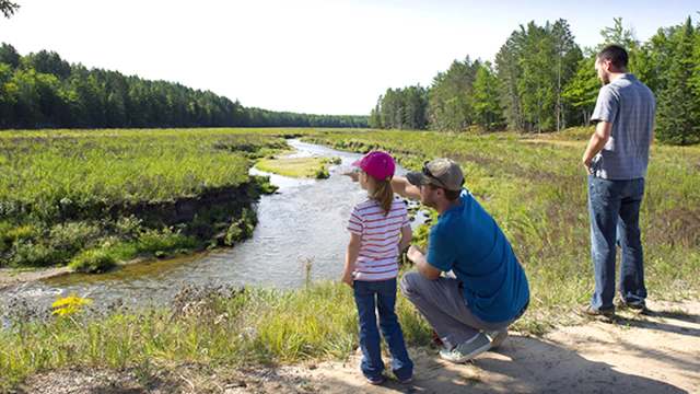 A child wearing a pink hat points at a river while an adult crouches beside her and another adult stands nearby.