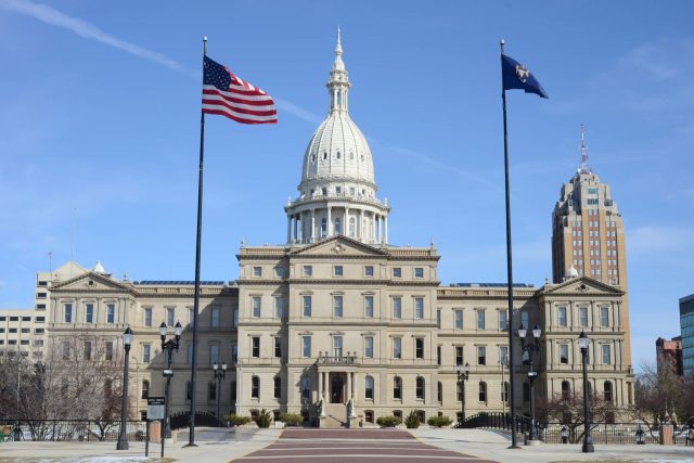 The Michigan State Capitol building with flags flying, surrounded by a clear blue sky.