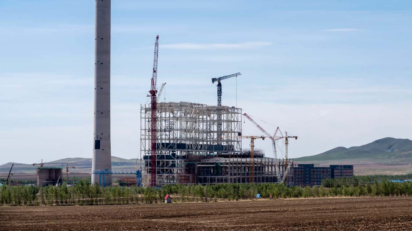 A large industrial construction site with cranes and a tall chimney against a mountainous backdrop.