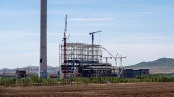 A large industrial construction site with cranes and a tall chimney against a mountainous backdrop.