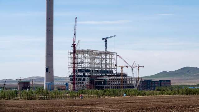 A large industrial construction site with cranes and a tall chimney against a mountainous backdrop.