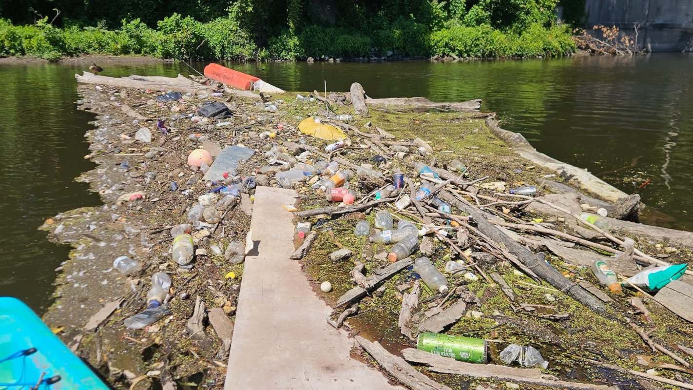 A large pile of plastic waste and debris floating in a river, surrounded by greenery.