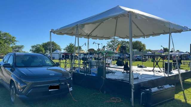 A black SUV parked next to a covered stage with musical instruments and a clear blue sky in the background.