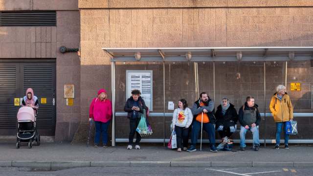 A diverse group of people waiting at a bus stop, some engaging on phones and others chatting.