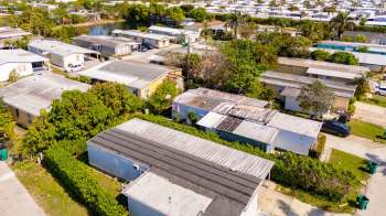 An aerial view of a residential area featuring mobile homes, greenery, and a small pond in the background.