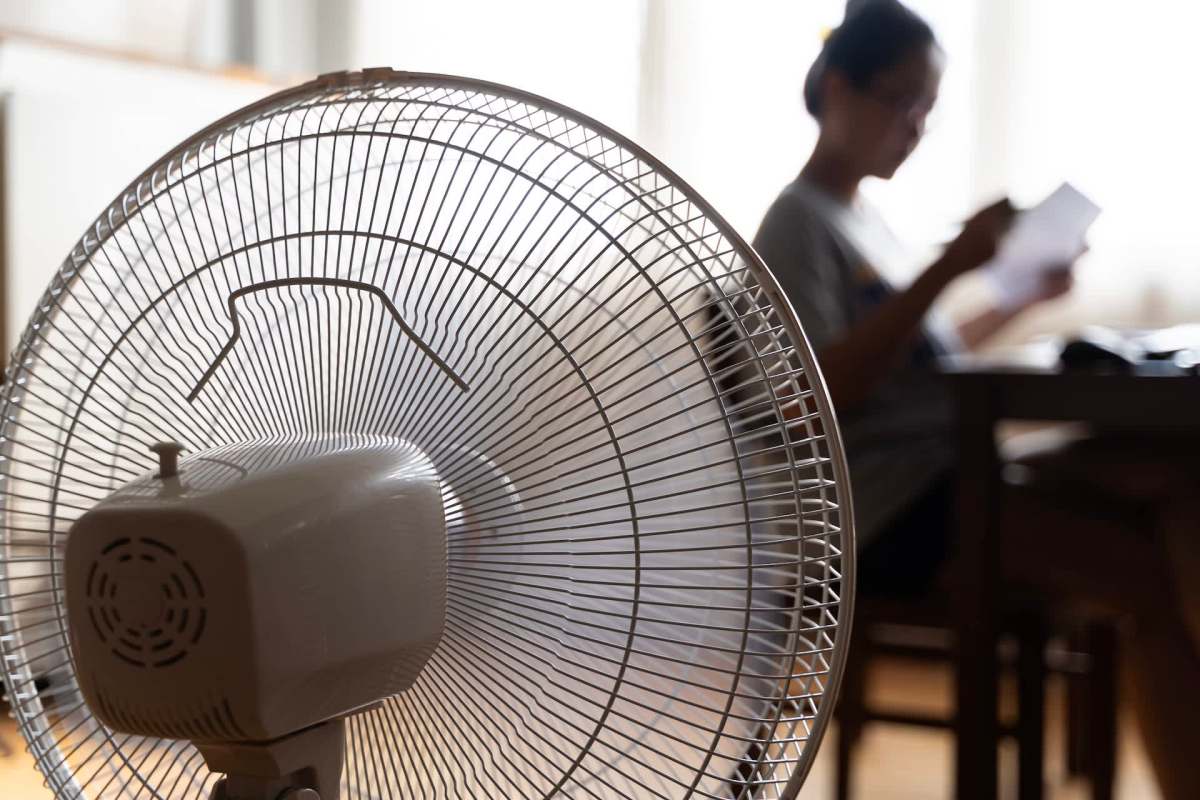 A close-up of an electric fan in the foreground with a person reading in the background.