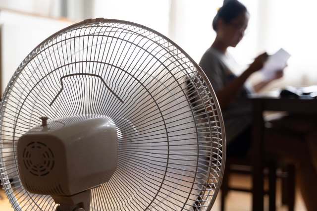 A close-up of an electric fan in the foreground with a person reading in the background.
