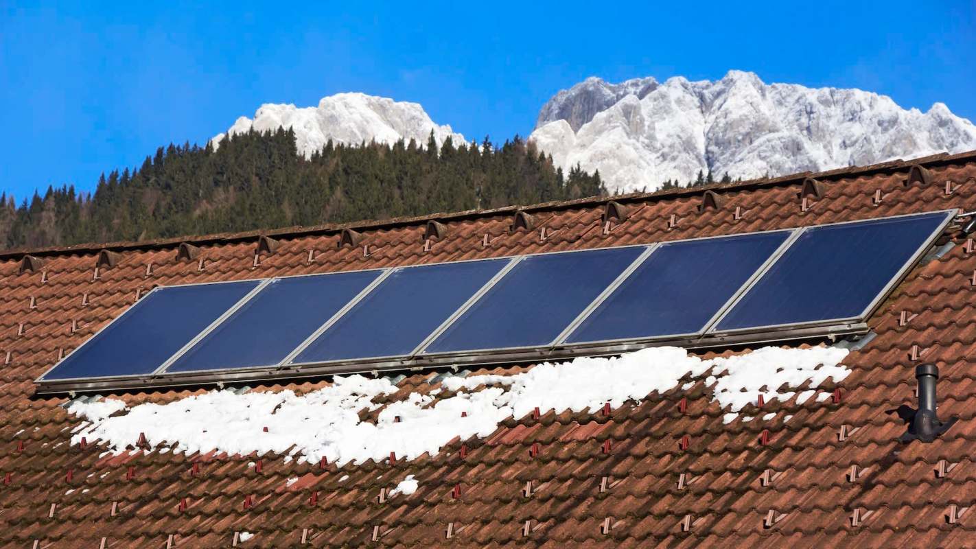 Solar panels on a snowy rooftop with mountains in the background under a clear blue sky.