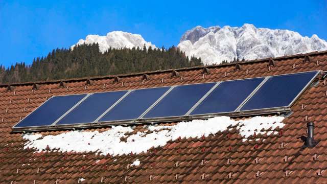 Solar panels on a snowy rooftop with mountains in the background under a clear blue sky.