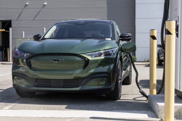A green Ford Mustang Mach-E charging at a station with an industrial backdrop.