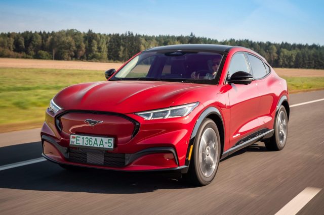 A red electric SUV is driving on a rural road surrounded by a green landscape under a clear blue sky.