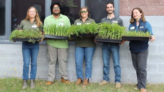 Five individuals stand together holding trays of plants in front of a building, smiling and wearing matching shirts.