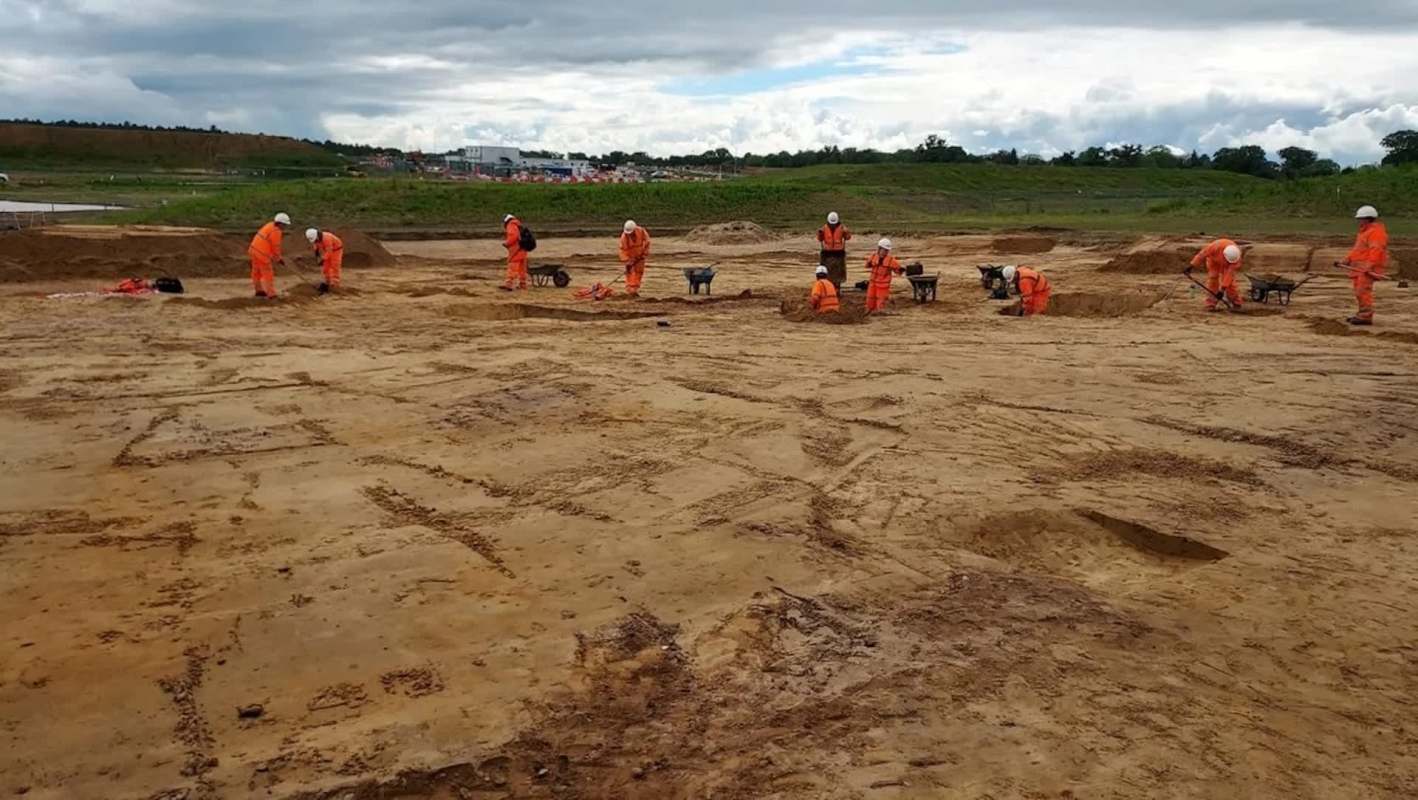 Workers in orange uniforms and helmets excavate a sandy brown dirt site under a cloudy sky.