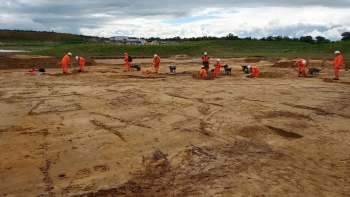 Workers in orange uniforms and helmets excavate a sandy brown dirt site under a cloudy sky.