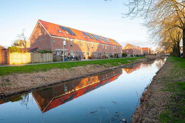 Residential buildings with red rooftops reflected in a calm canal, lined by trees and grassy banks.