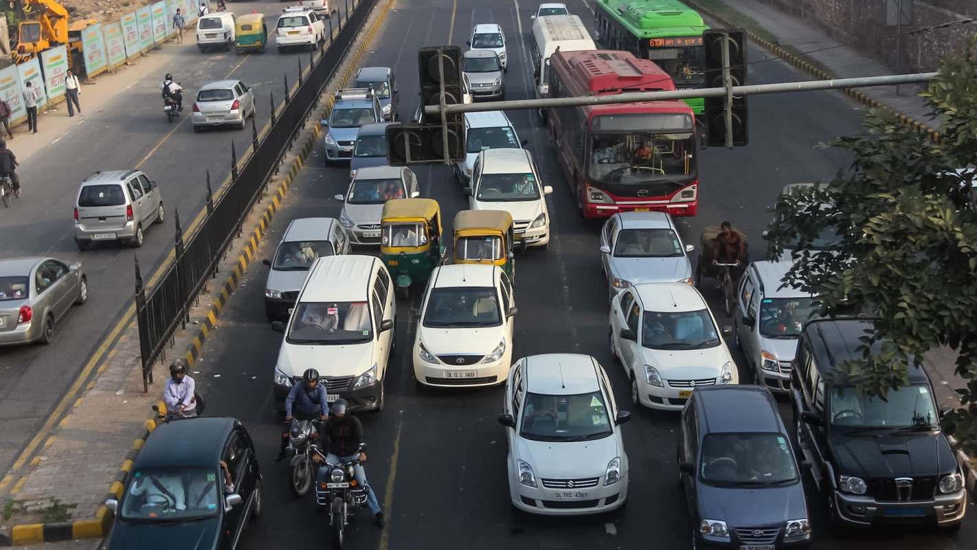 A busy urban road with congested traffic, featuring cars, auto-rickshaws, and a red bus at a traffic signal.