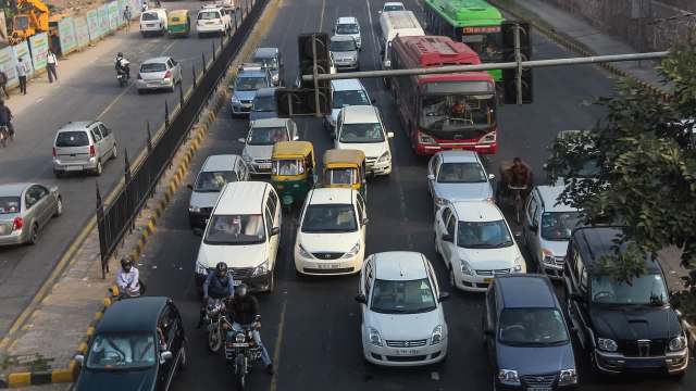 A busy urban road with congested traffic, featuring cars, auto-rickshaws, and a red bus at a traffic signal.