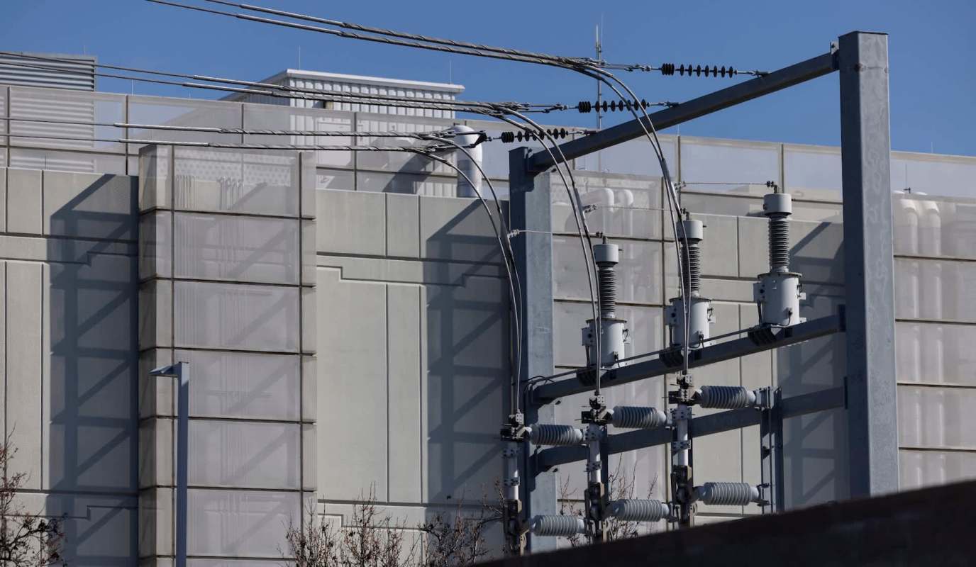 Power lines and electrical equipment are positioned against a modern building with a clear blue sky in the background.