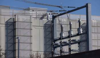Power lines and electrical equipment are positioned against a modern building with a clear blue sky in the background.