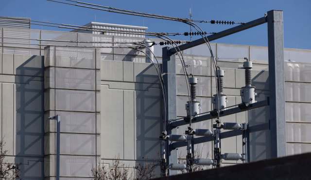 Power lines and electrical equipment are positioned against a modern building with a clear blue sky in the background.