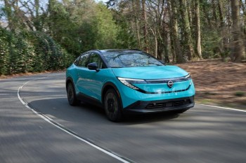 A teal electric SUV drives along a winding road surrounded by lush greenery and trees.