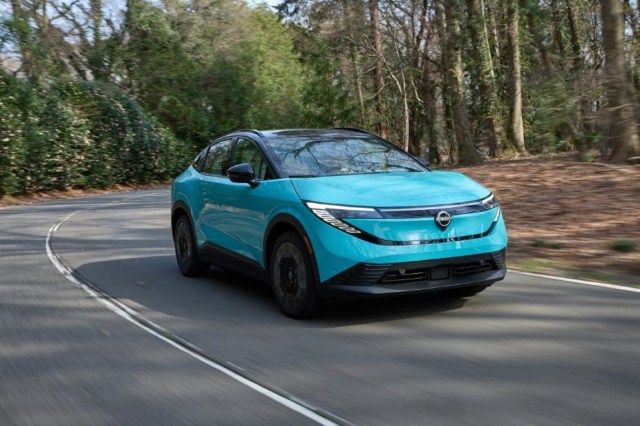 A teal electric SUV drives along a winding road surrounded by lush greenery and trees.