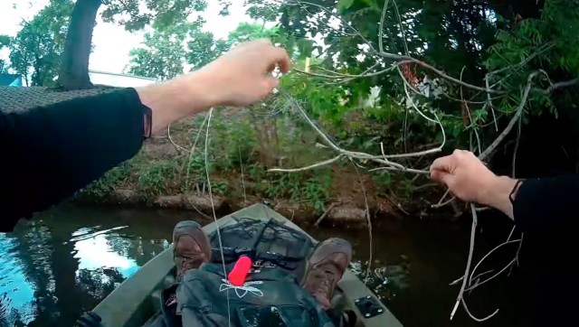 A person in a kayak is untangling fishing lines from branches overhanging a calm waterway.