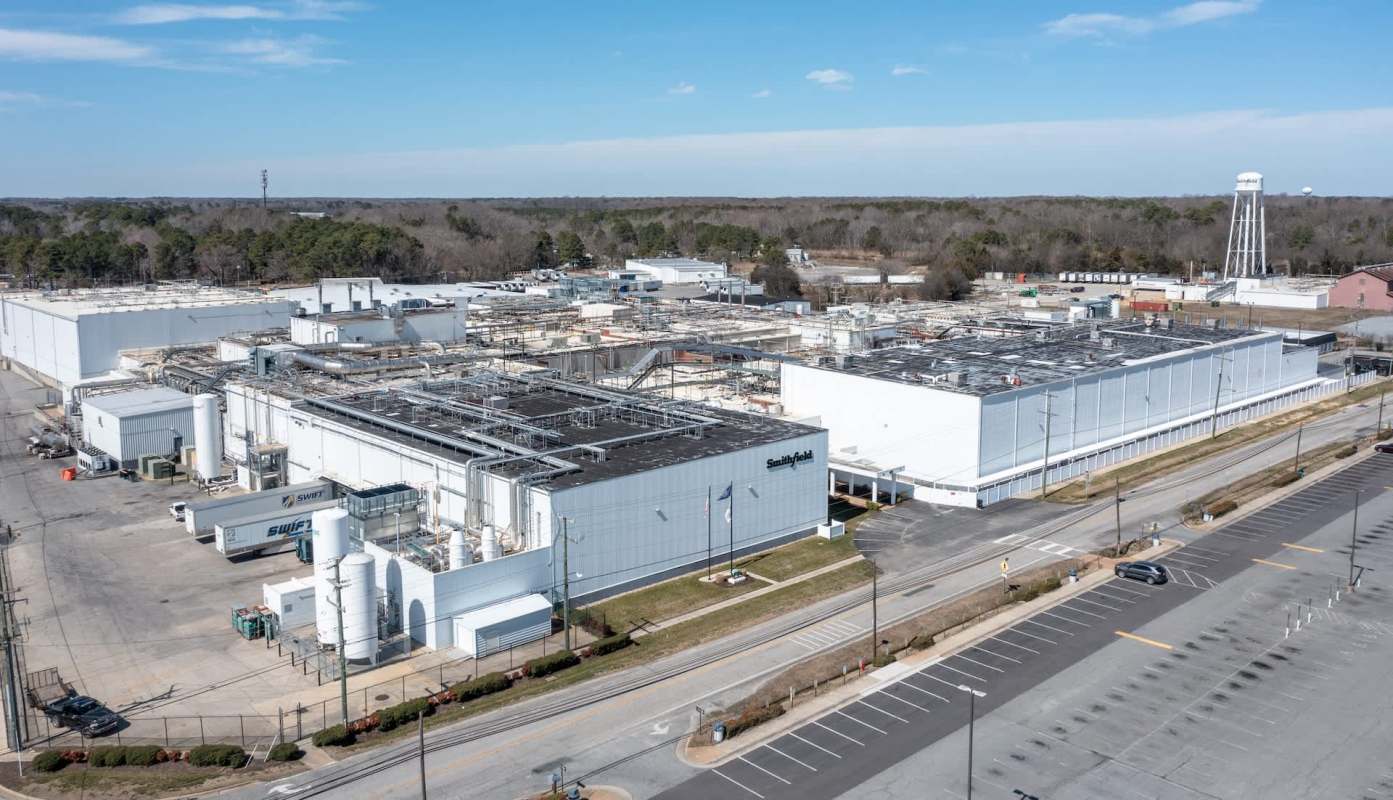 An industrial facility with multiple buildings and equipment, located near wooded areas and an empty parking lot.