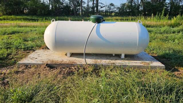 A white propane tank sits on a concrete pad surrounded by tall grass and trees in a rural area.