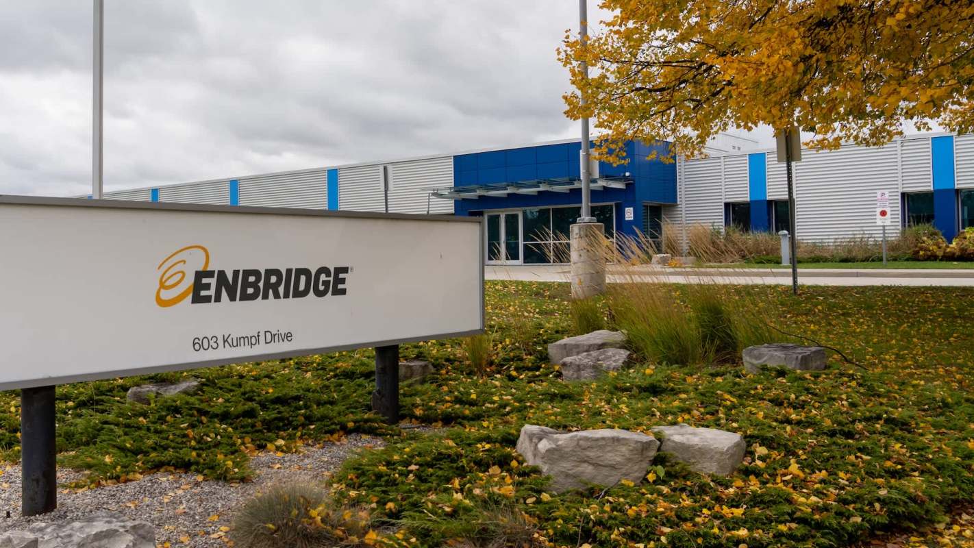 A sign for Enbridge at 603 Kumpf Drive, with an autumn landscape and a modern building in the background.