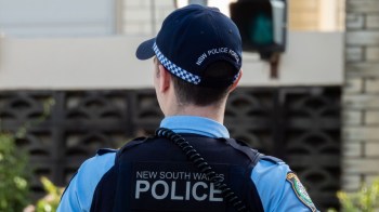 A police officer in uniform stands with their back to the camera, wearing a cap labeled "NSW Police Force."