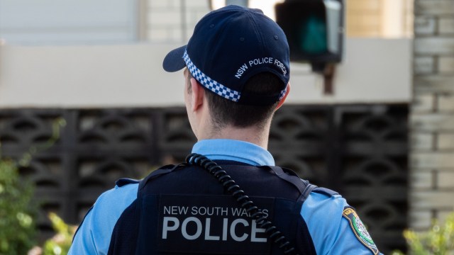 A police officer in uniform stands with their back to the camera, wearing a cap labeled "NSW Police Force."