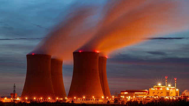 Three cooling towers emit steam into the twilight sky, with lights illuminating a nearby power plant.