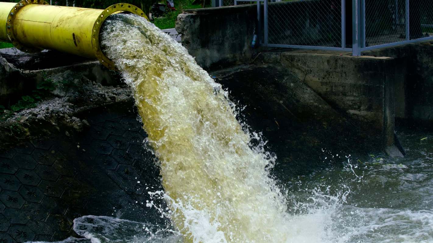 A yellow pipe discharges murky water into a pond, creating splashes and ripples on the surface.