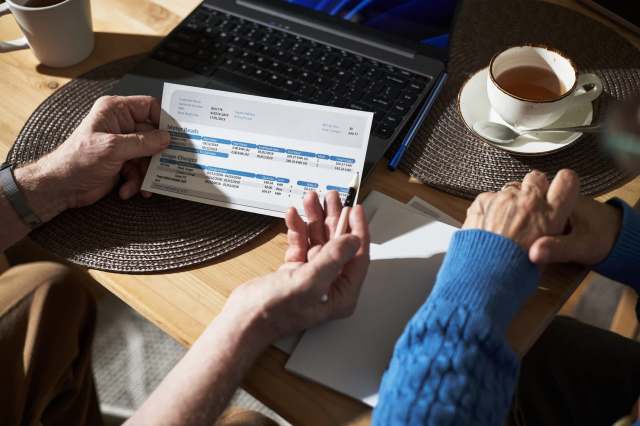 Two elderly hands holding a bill while a laptop and a cup of tea are visible on a wooden table.
