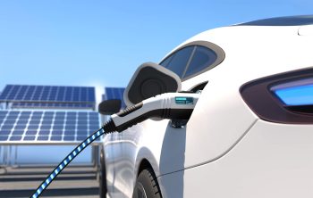 An electric car is plugged into a charging station near solar panels under a clear blue sky.