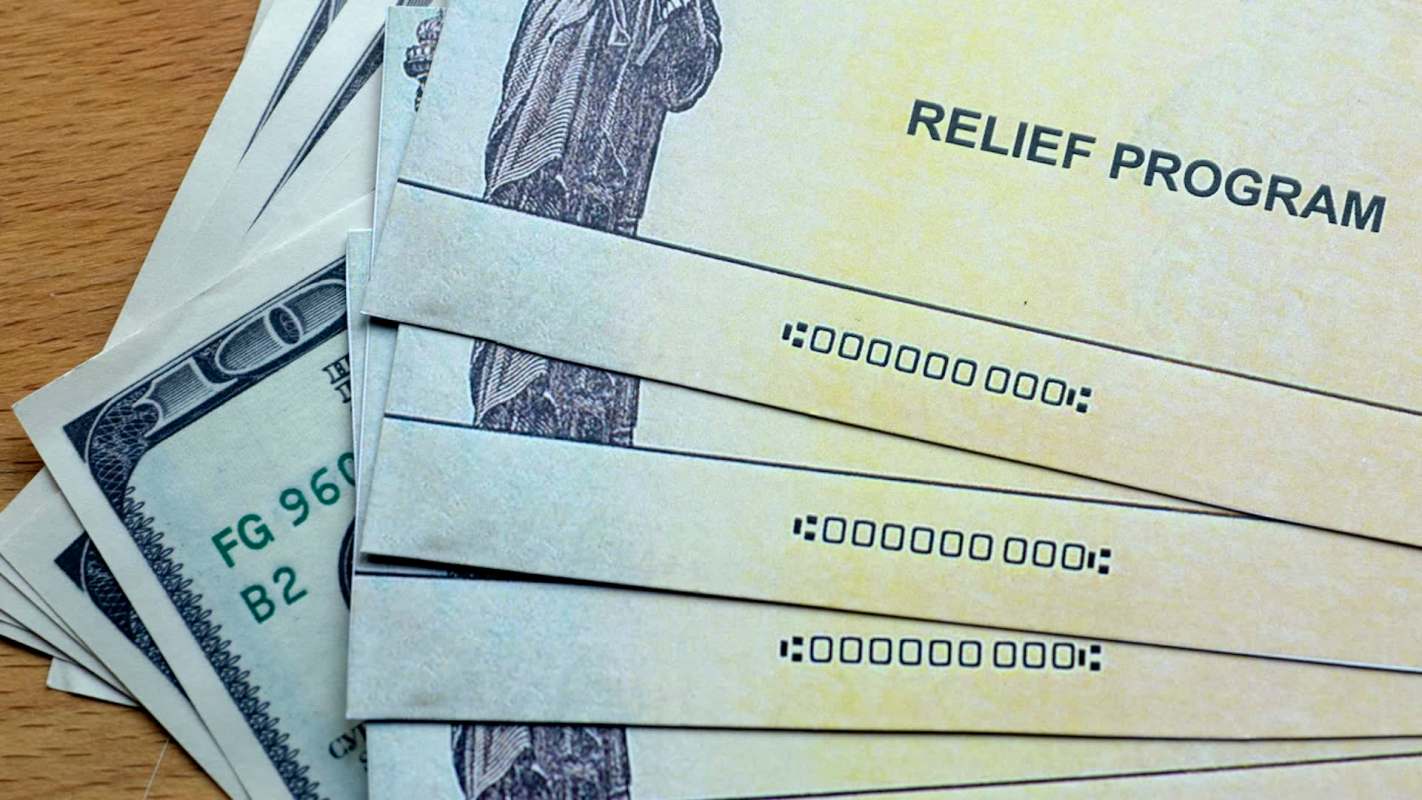 A close-up of relief program checks stacked next to a few $100 bills on a wooden surface.