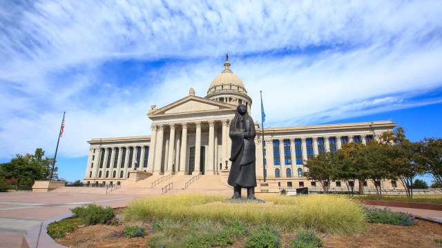 A bronze statue of a woman stands in front of a large government building under a bright blue sky.