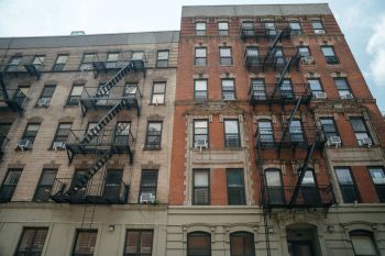 A close-up view of a red-brick apartment building featuring black fire escapes and air conditioning units.
