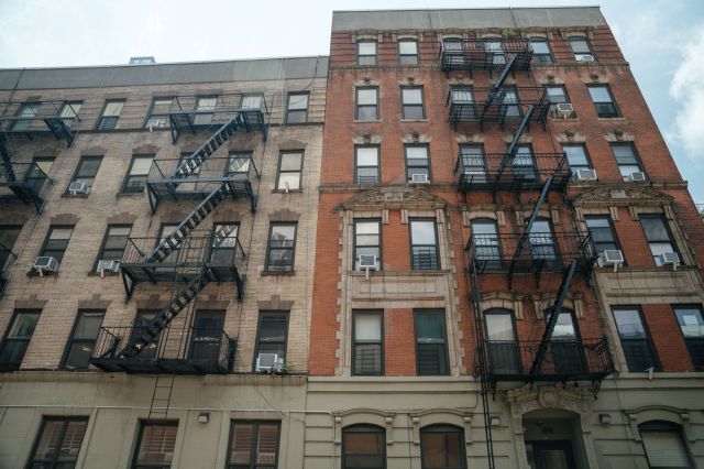 A close-up view of a red-brick apartment building featuring black fire escapes and air conditioning units.