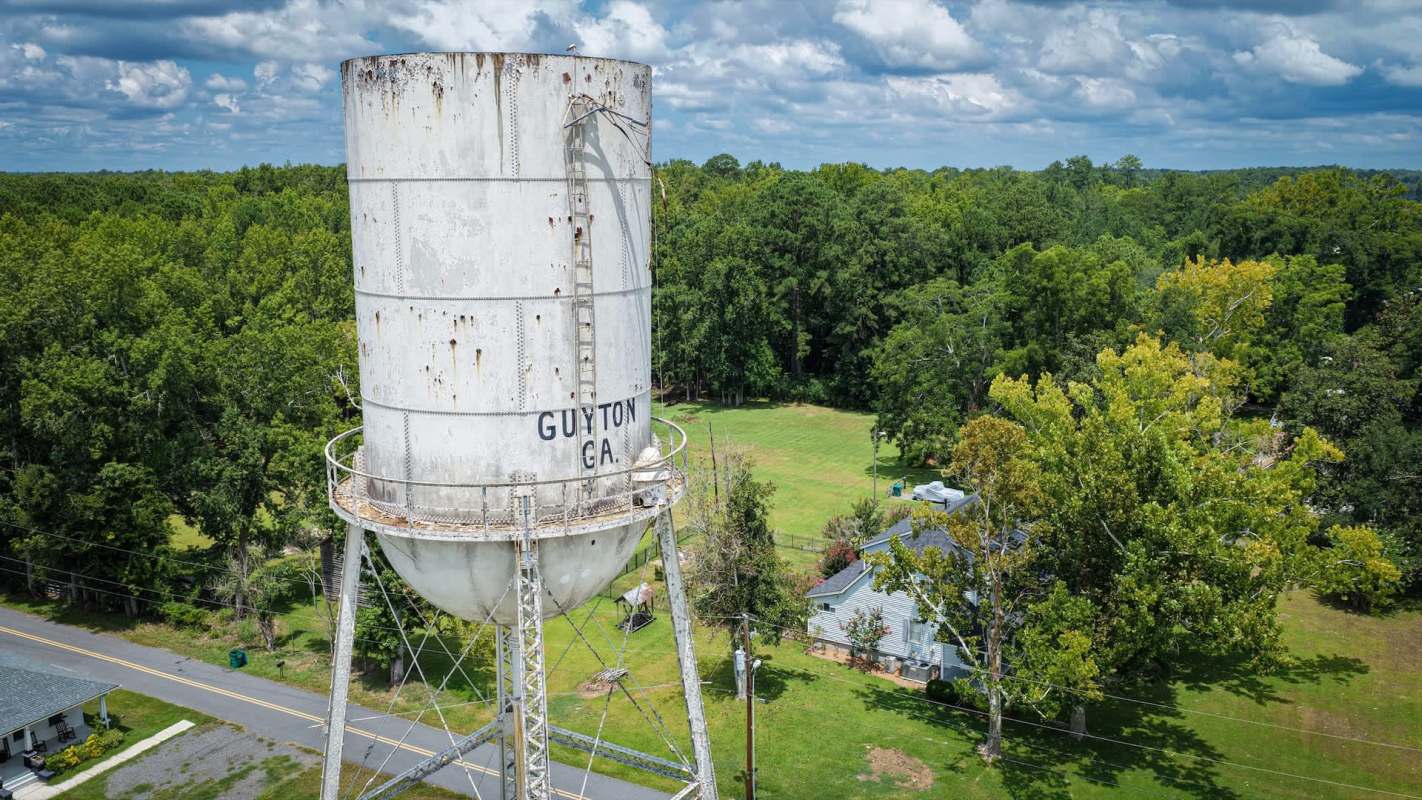 A water tower with the text "GUYTON GA" surrounded by green trees and a house nearby.